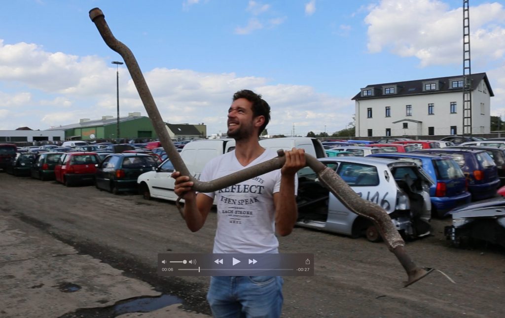 Ausgebaute, verrostete Auspuffanlage des T4, hochgehalten auf einem Parkplatz