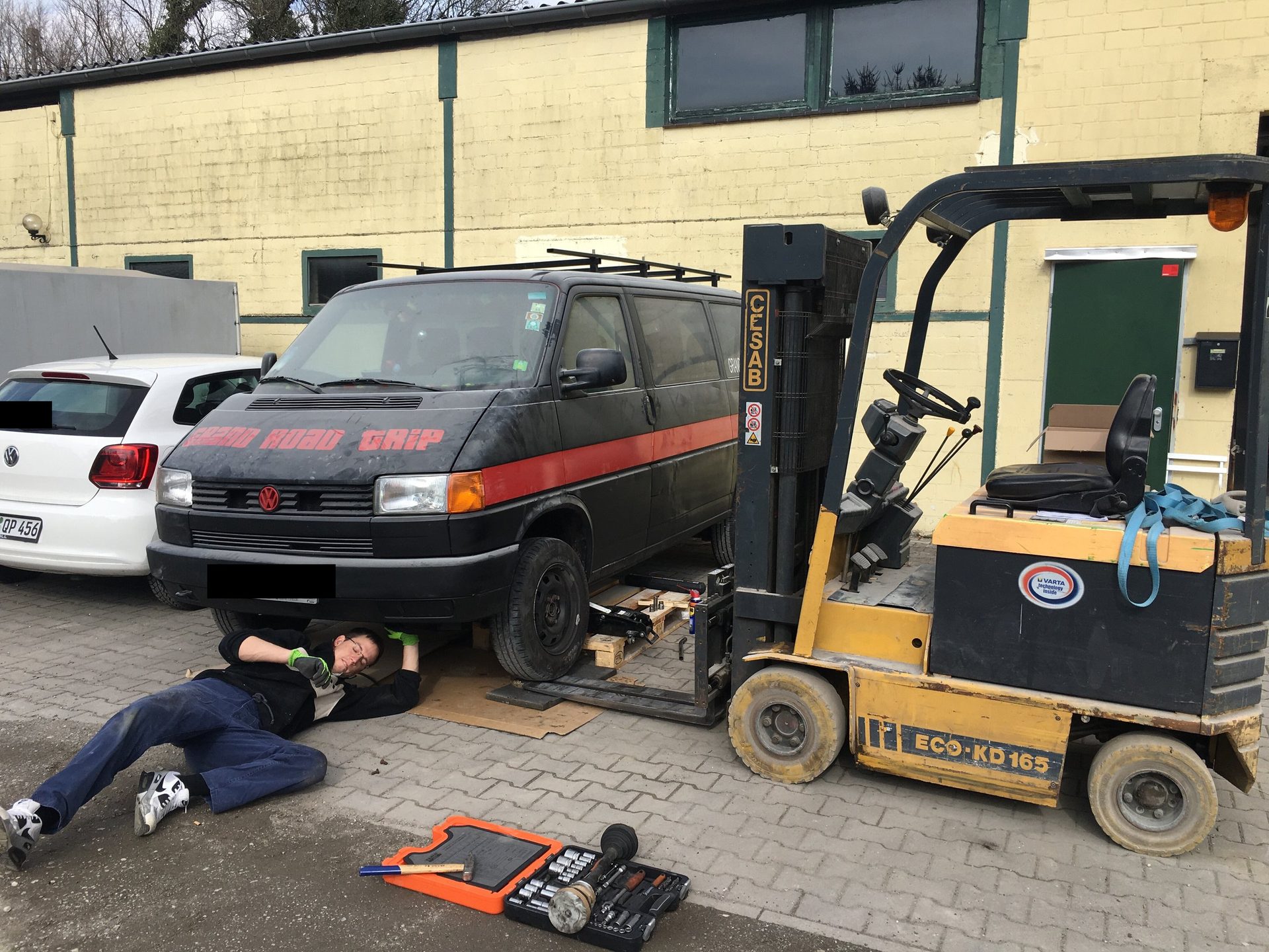 Der aufgebockte T4, daneben ein Gabelstapler, darunter liegt jemand mit Werkzeug